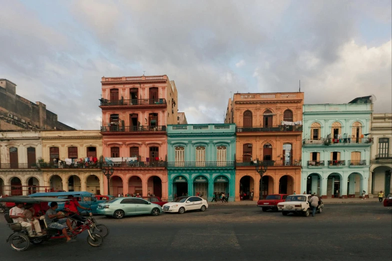 a group of cars driving down a street next to tall buildings, a colorized photo, by Ceferí Olivé, pexels contest winner, hyperrealism, colorful houses, cuban setting, square, late afternoon