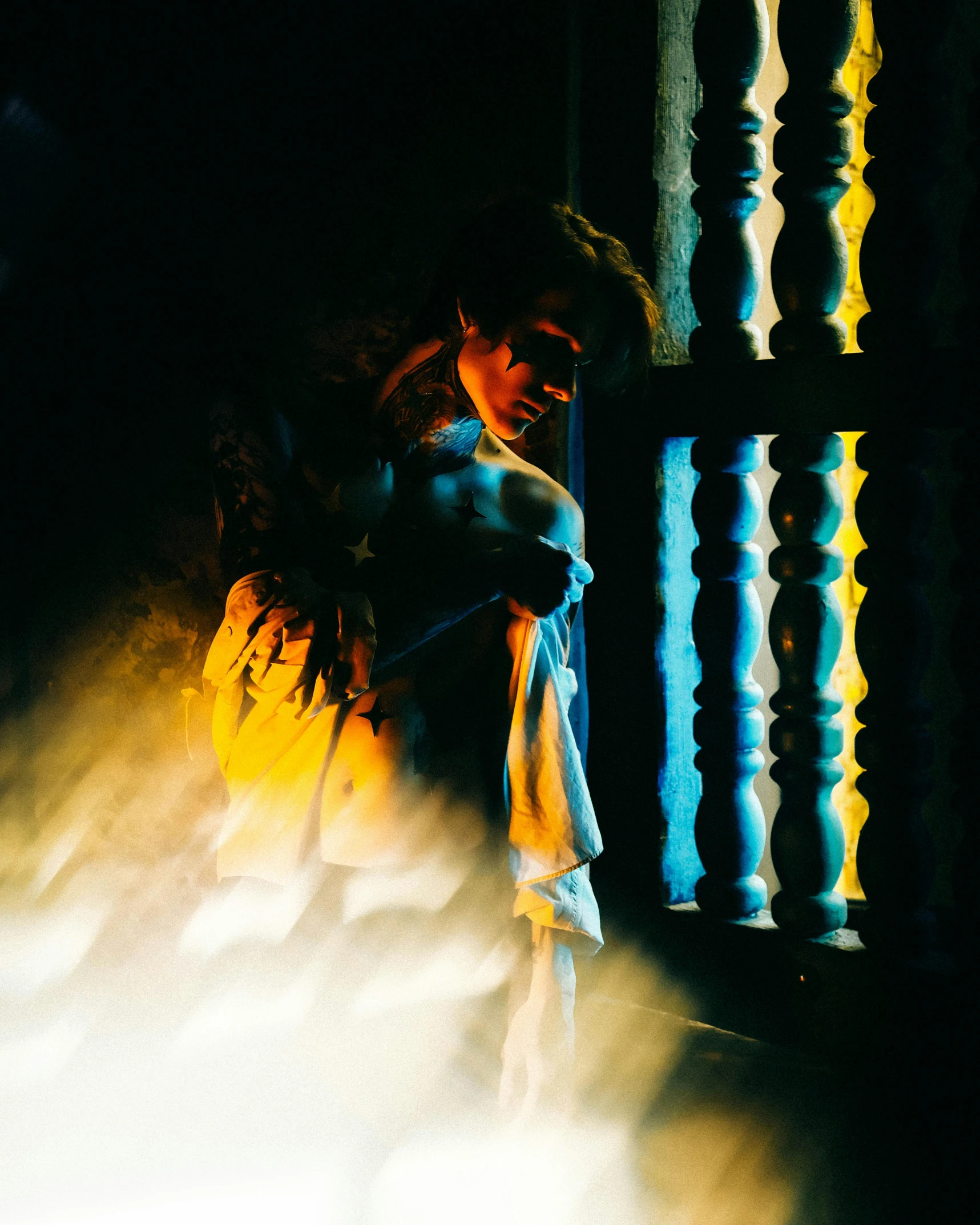 a woman leaning against a railing in the dark, an album cover, pexels contest winner, romanticism, smoke filled room, light over boy, blue and yellow lighting, clad in robes