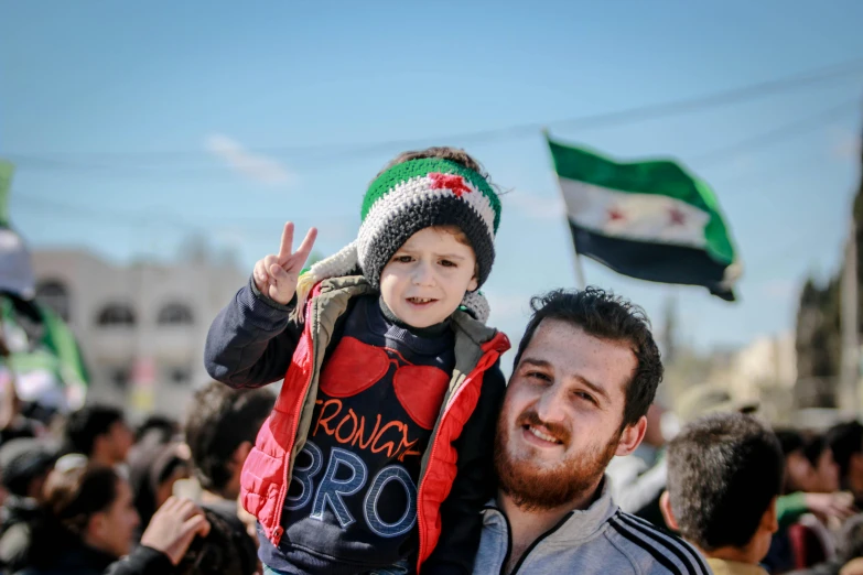 a man holding a child in front of a crowd of people, pexels contest winner, hurufiyya, balaclava, square, revolutionary, february)