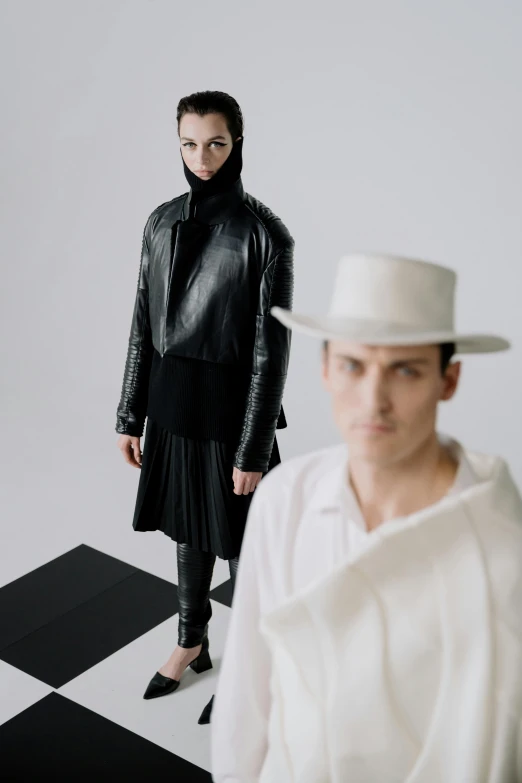 a man and a woman standing in front of a black and white checkered floor, an album cover, inspired by Marina Abramović, trending on unsplash, bauhaus, tall hat, nun fashion model, leather and suede, on a white table