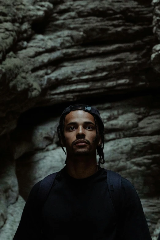 a man standing in front of a rock formation, dark background, light skin, ariel perez, drake