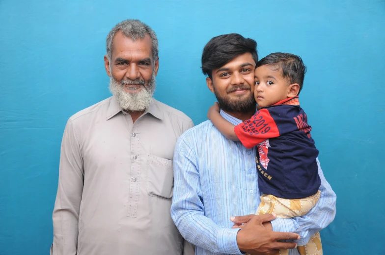 a man holding a child in front of a blue wall, by Riza Abbasi, without beard and mustache, group photo, 15081959 21121991 01012000 4k, customers