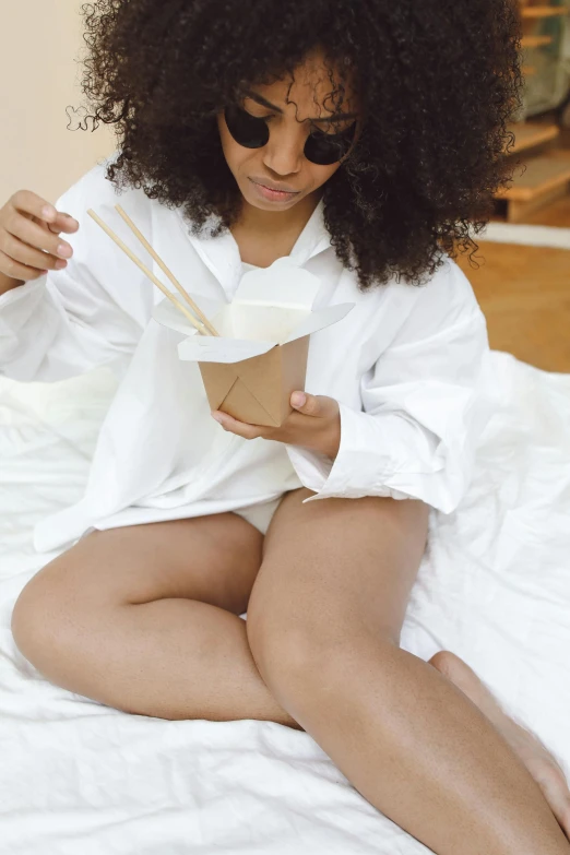 a woman sitting on top of a bed holding chopsticks, wearing white pajamas, eating noodles, tan skin a tee shirt and shorts, 2019 trending photo