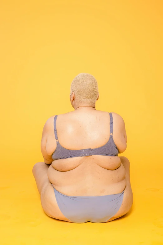 an overweight woman sitting on a yellow surface, by Ren Hang, trending on pexels, in a black betch bra, bald lines, lower back, grey skinned