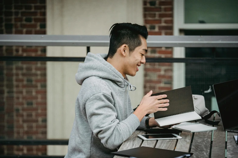 a man sitting at a table using a laptop computer, pexels contest winner, academic art, waving and smiling, wearing a grey hooded sweatshirt, holding books, asian man