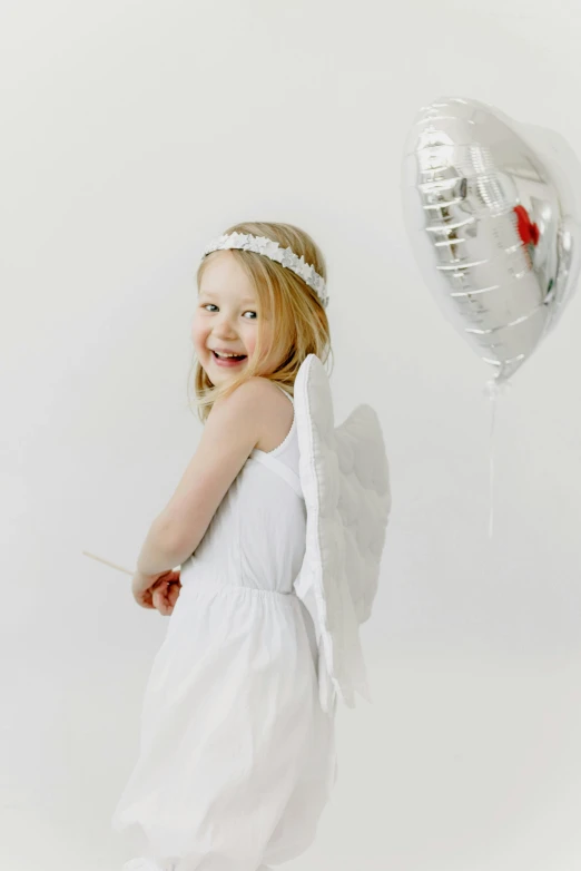 a little girl dressed as an angel holding a heart balloon, silver angel wings, wearing a paper crown, full product shot, white