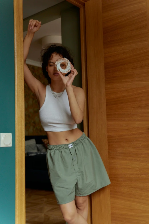 a woman standing in a doorway holding a donut, inspired by Elsa Bleda, trending on pexels, posing in bed, croptop, muted green, kiko mizuhara