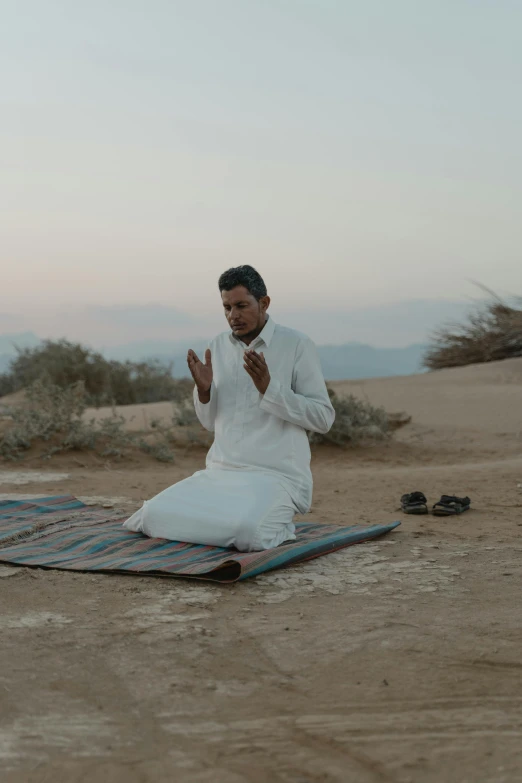 a man sitting on a blanket in the desert, an album cover, inspired by Kamāl ud-Dīn Behzād, unsplash, hurufiyya, praying posture, movie still frame, performing a music video, telling stories