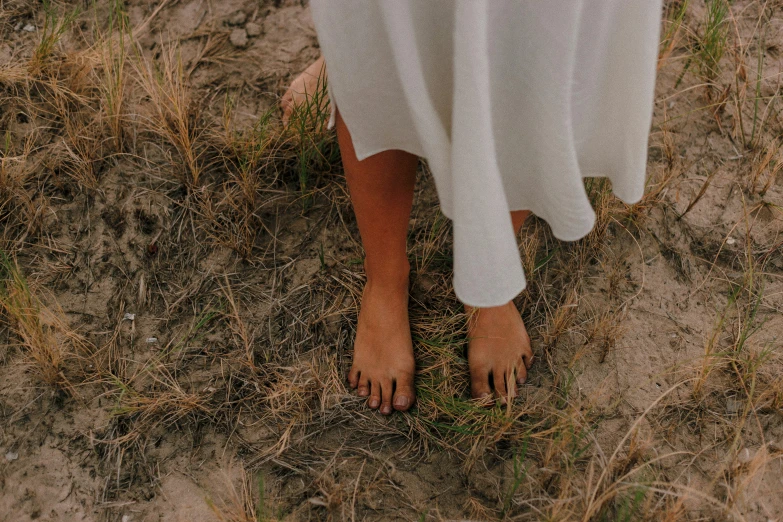 a woman in a white dress standing in a field, unsplash, bare feet in grass, brown skin like soil, standing on rocky ground, visible pores