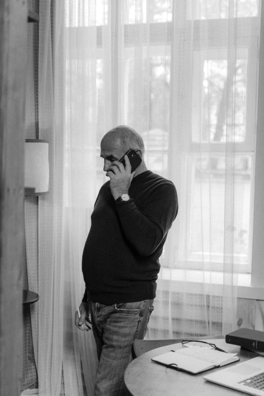 a man standing in front of a window talking on a cell phone, a black and white photo, by Edi Rama, bald man, vladimir krisetskiy, middle - age, at home