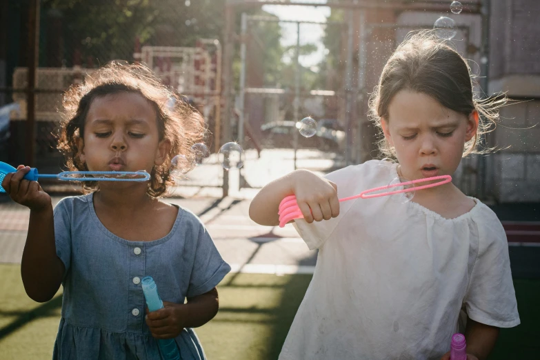 a couple of young girls standing next to each other, by Lee Loughridge, pexels contest winner, spit flying from mouth, soap bubble mind, urban playground, young girl playing flute