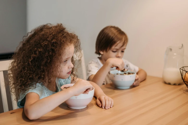 two little girls sitting at a table eating cereal, by Emma Andijewska, pexels contest winner, boy and girl, avatar image, looking left, bowl