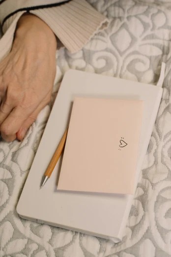 a person writing on a piece of paper on a bed, rose gold heart, holding notebook, wearing a light - pink suit, uploaded