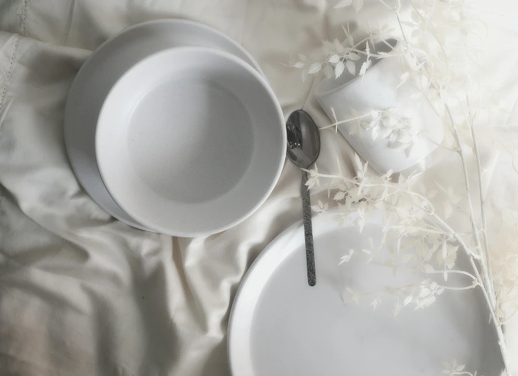 a white plate sitting on top of a bed next to a cup and saucer, an ambient occlusion render, inspired by Cerith Wyn Evans, unsplash, white splendid fabric, cutlery, background image