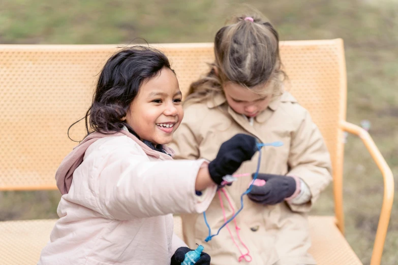 a couple of little girls sitting on top of a bench, wearing gloves, wrapped in wires and piones, casual playrix games, manuka