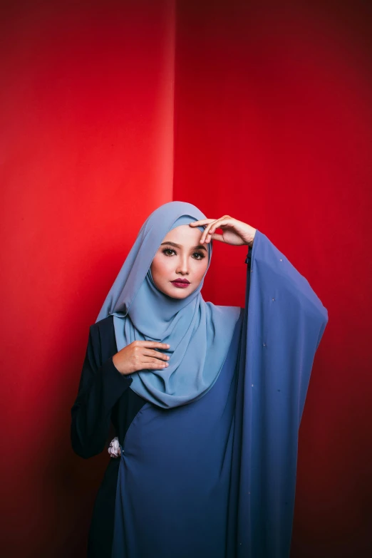 a woman in a blue hijab standing against a red wall, an album cover, by Basuki Abdullah, trending on pexels, blue gray, 5 0 0 px models, shawl, pose 4 of 1 6