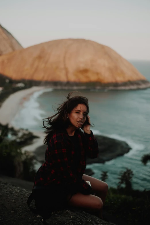 a woman sitting on top of a hill next to the ocean, inspired by Elsa Bleda, trending on pexels, happening, caramel. rugged, flannel, detailed unblurred face, her hair flowing down