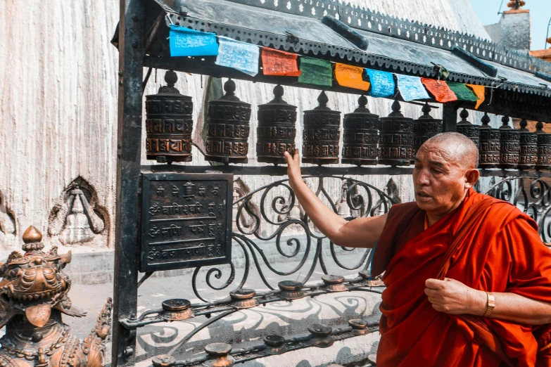 a man in a red robe standing next to a wall with bells, by Sam Dillemans, pexels contest winner, cloisonnism, thumbnail, lama, temples behind her, brown