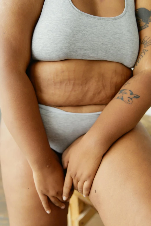 a woman sitting on top of a wooden chair, by Matija Jama, trending on pexels, renaissance, her belly is fat and round, big symmetrical scar features, tattoos all over the skin, failed cosmetic surgery