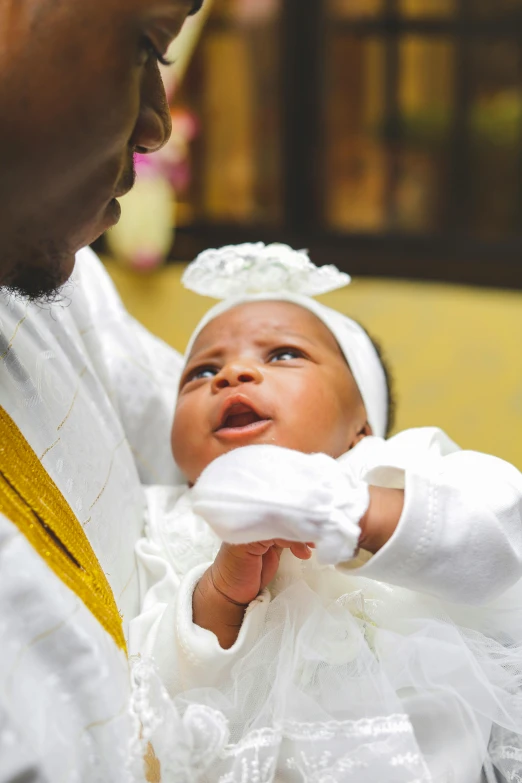 a man holding a baby in a white dress, an album cover, by Chinwe Chukwuogo-Roy, pexels, baptism, white and gold color scheme, holy ceremony, little kid