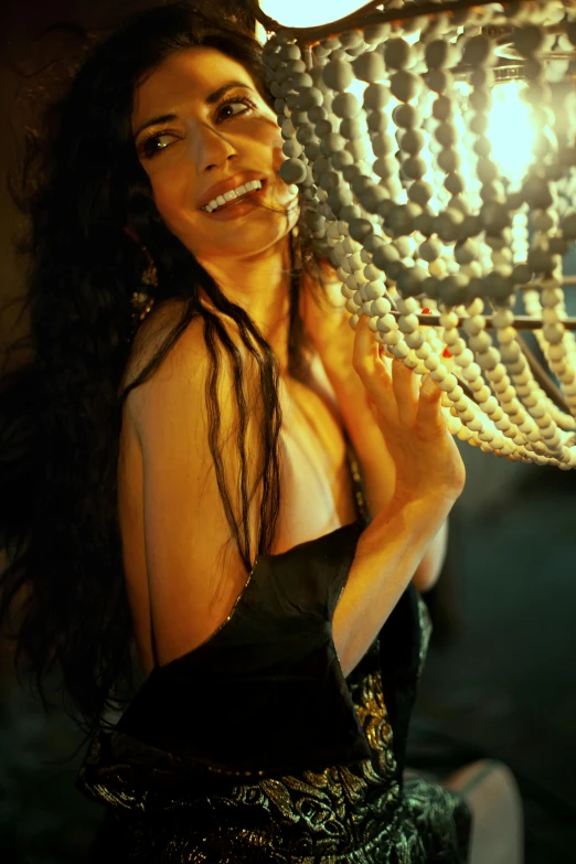 a woman in a black dress holding a chandelier, long black curly hair, smiling seductively, back lighting, film still promotional image