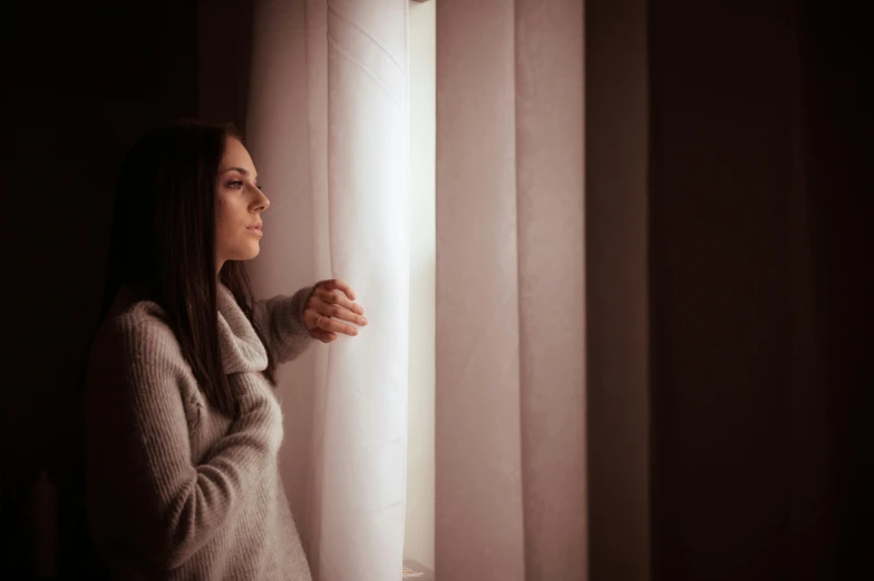 a woman looking out a window in a dark room, a picture, white light shining on her, autumn season, thoughtful, at home