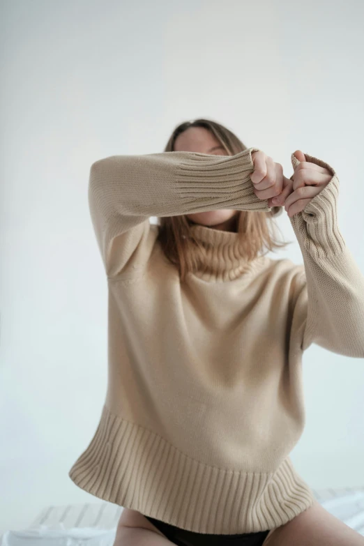 a woman sitting on top of a bed covering her eyes, by Helen Stevenson, trending on pexels, wearing turtleneck, beige, made of wool, playful pose