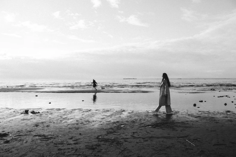 a woman standing on top of a beach next to the ocean, a black and white photo, by Caro Niederer, unsplash, children playing at the beach, walking on water, cinematic. by leng jun, oyasumi punpun