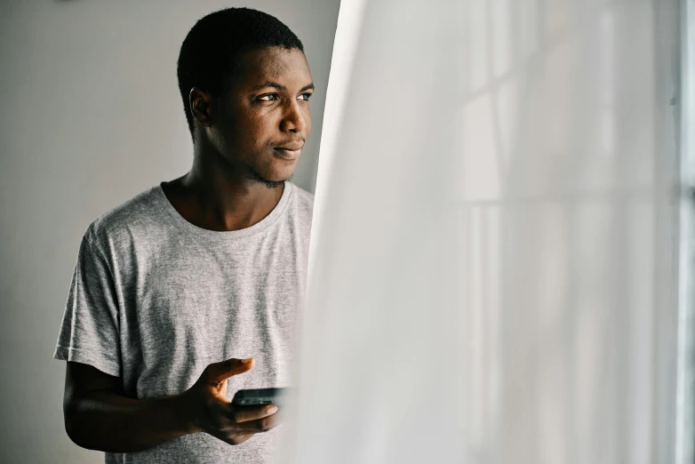 a man standing in front of a window holding a cell phone, black man, wearing a light shirt, slightly minimal, wearing translucent sheet