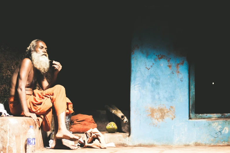 a man sitting in front of a blue building, inspired by Steve McCurry, pexels contest winner, bengal school of art, praying with tobacco, cottage hippie naturalist, an oldman, thumbnail