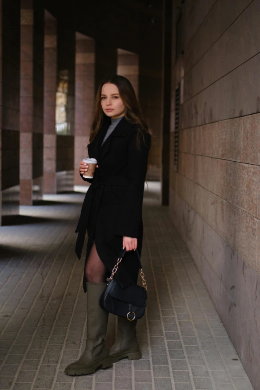 a woman standing next to a wall holding a cup of coffee, by Emma Andijewska, lady in black coat and pantyhose, at college, dasha taran, professional image