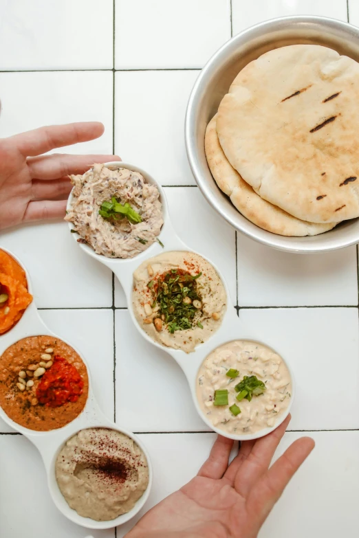 a table topped with bowls of different types of food, dau-al-set, sleek hands, humus, 6 pack, round-cropped