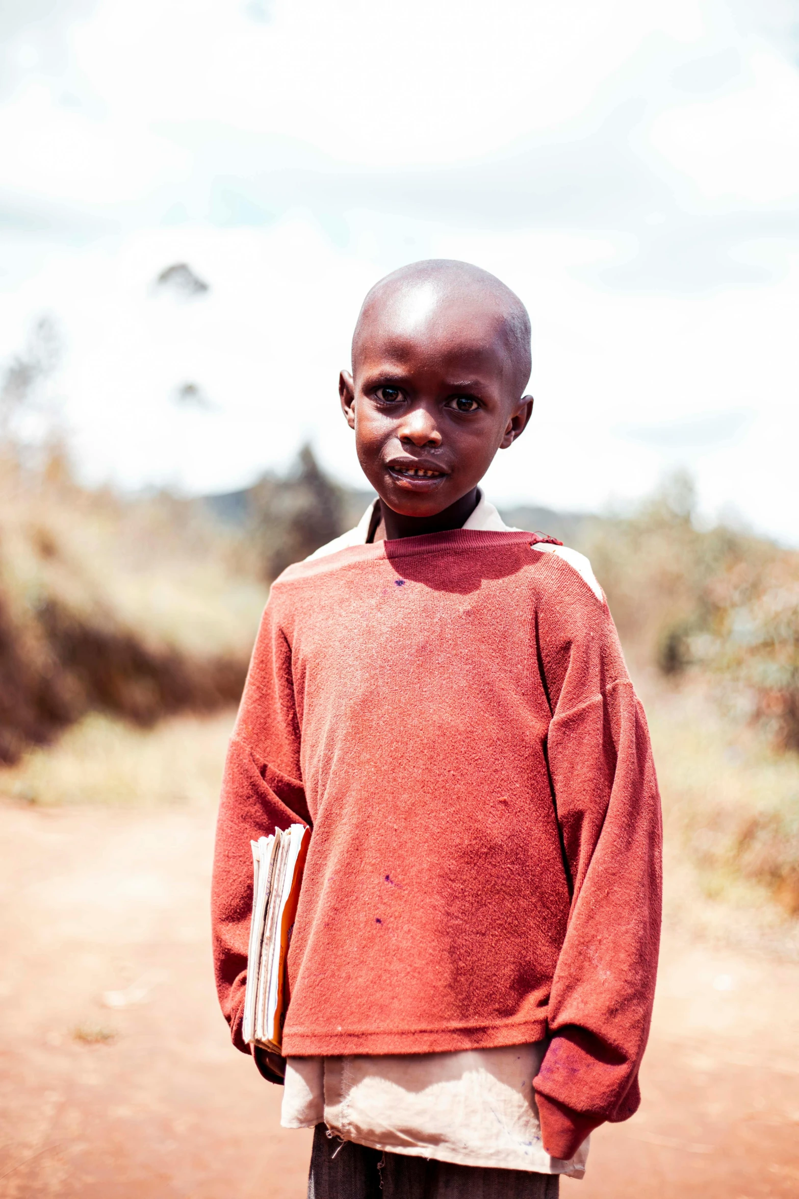 a young boy standing on a dirt road, very kenyan, holding notebook, fully dressed, at a clearing