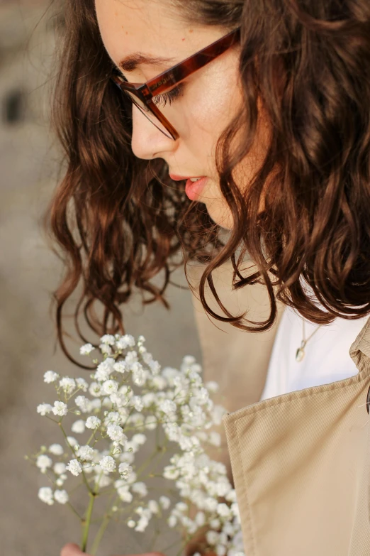 a woman holding a bunch of baby's breath flowers, inspired by Elsa Bleda, trending on unsplash, romanticism, wearing a long beige trench coat, curly brown hair, wavy long black hair and glasses, portrait of timothee chalamet