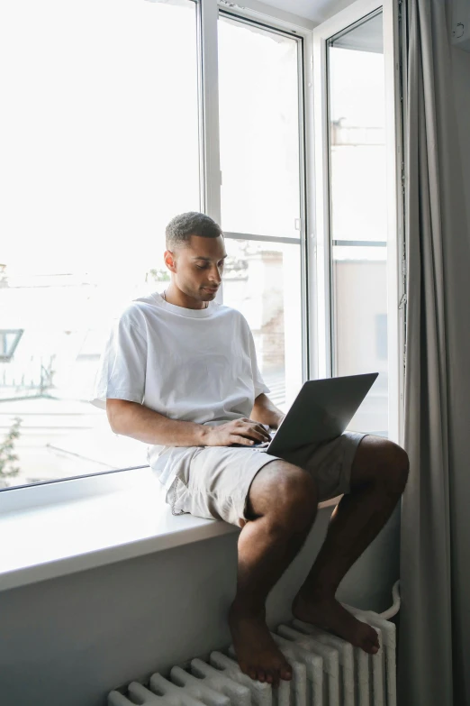 a man sitting on a window sill using a laptop, by Carey Morris, pexels contest winner, wearing shorts and t shirt, wearing a white button up shirt, black man, sitting in a lounge