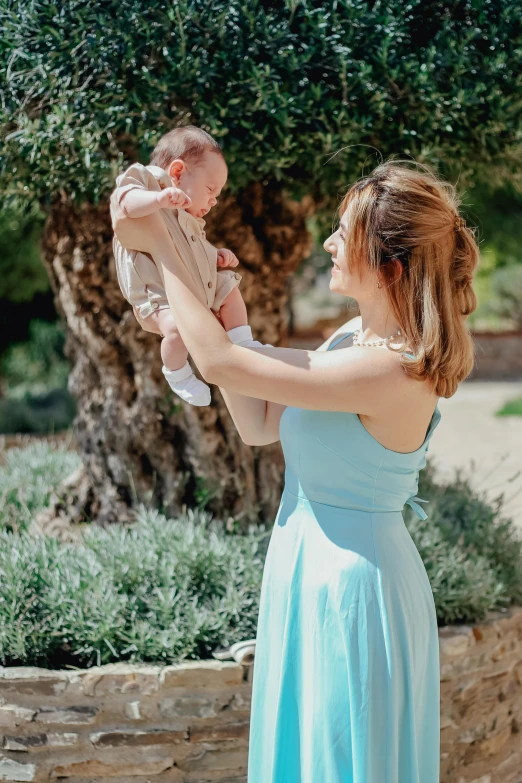 a woman in a blue dress holding a baby, by Romain brook, pexels contest winner, in a garden, soft silk dress, sleeveless, in spain