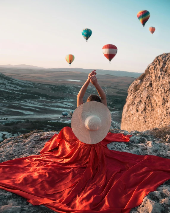 a woman in a red dress and hat sitting on a rock with hot air balloons in the background, pexels contest winner, flowing white robes, instagram post, rainbow clothes, cinematic outfit photo