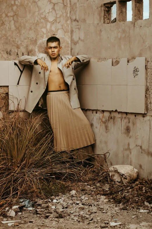 a man in a trench coat leaning against a wall, an album cover, inspired by Carlos Berlanga, trending on pexels, renaissance, white skirt and barechest, fashion shoot 8k, asian male, brutalist fashion show