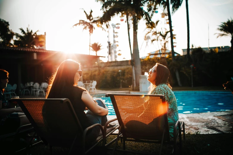 a couple of women sitting next to a swimming pool, by Lee Loughridge, pexels contest winner, evening sunlight, afternoon hangout, hotel room, back yard