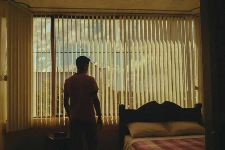 a man standing in front of a window next to a bed, inspired by Elsa Bleda, unsplash, hyperrealism, vhs footage still, moody : : wes anderson, curtain, blind