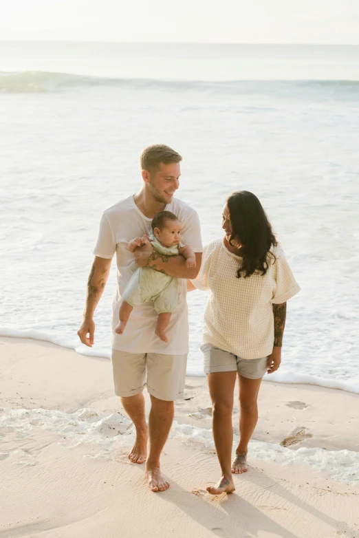 a man and woman walking on a beach with a baby, tan skin a tee shirt and shorts, white, liam, 2 people