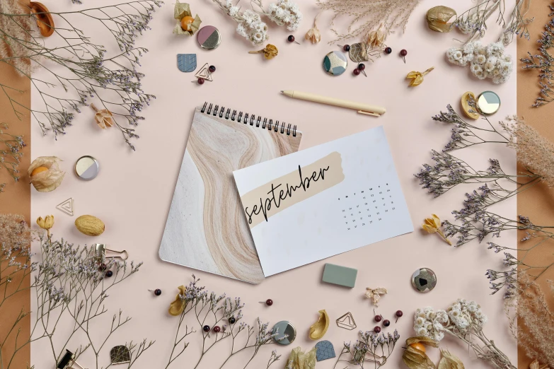 a calendar sitting on top of a table surrounded by dried plants, inspired by Eden Box, unsplash, happening, pastel pink concrete, striking pose, paper quilling, autumn colours