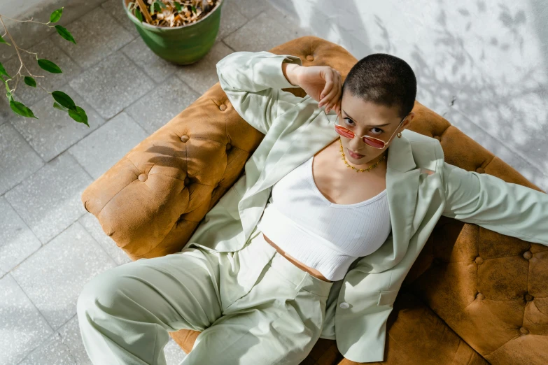 a woman sitting on top of a brown couch, by Emma Andijewska, trending on pexels, bright trouser suit for a rave, shaved hair, greenish skin, smooth white tight clothes suit