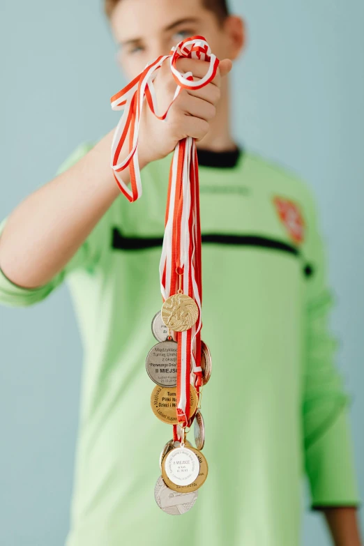 a young boy holding a bunch of medals, pexels contest winner, sports mascot, hanging, red ribbon, a green