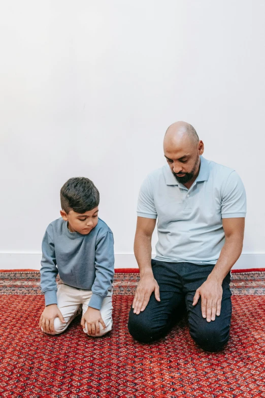 a man sitting on the floor next to a little boy, hurufiyya, kneeling in prayer, riyahd cassiem, father figure image