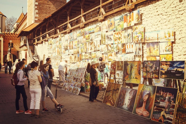 a group of people standing next to a wall with paintings on it, a picture, pexels contest winner, market stalls, sunny weather, brown, square