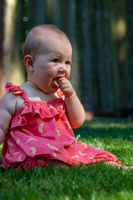 a baby sitting on top of a lush green field, wearing a red sundress, snacks, hand on her chin, organic dress