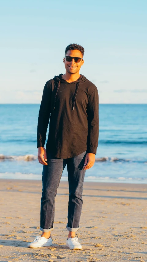 a man standing on a beach next to the ocean, wearing jeans and a black hoodie, wearing a kurta, thumbnail, manly