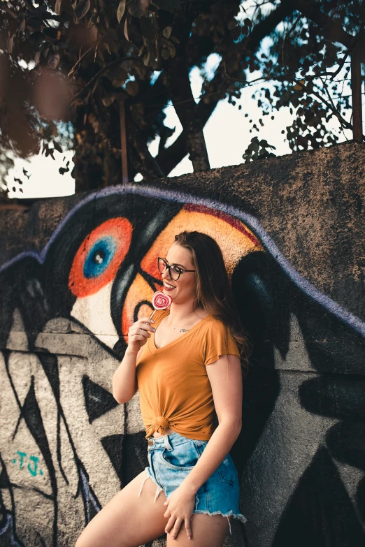 a woman standing next to a wall with graffiti on it, pexels contest winner, an ewok eating a lollipop, wearing big black circle glasses, wearing an orange t-shirt, flirting