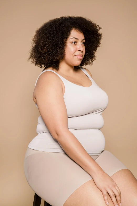 a woman sitting on a stool with her legs crossed, trending on pexels, big stomach, wearing a vest top, light-brown skin, obese )